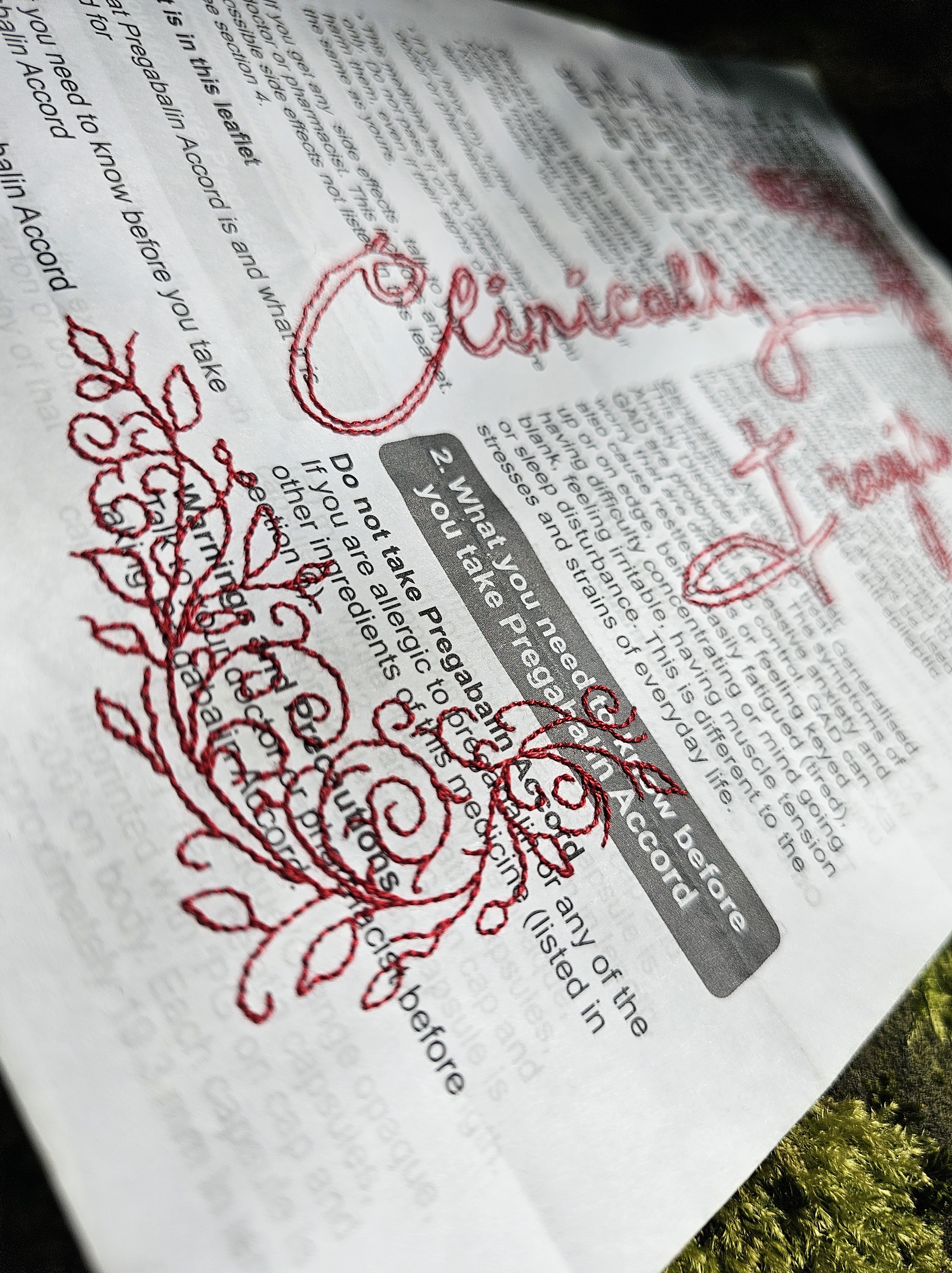 A close up of a medication packet with hand embroidered swirling vines and leaves that create a corner frame. These have been backstitched in red thread to stand out against the white of the leaflet.