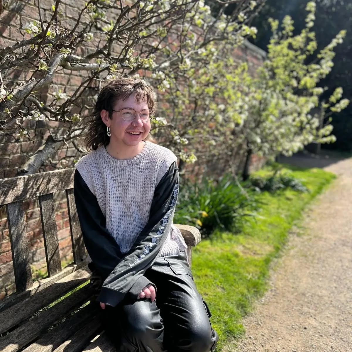 Finn - a white non-binary person with a brown wavy mullet, glasses and facial piercings - is sat on a bench, looking over to the right. They are wearing a handmade crochet vest in light grey, a black long-sleeved top with a barbed wire motif running down the arms, and black pleather trousers. In the background are blossoming plants against a brick wall, a grass verge, and a pebbled pathway. The weather is sunny, and Finn's hair is blowing in the wind.