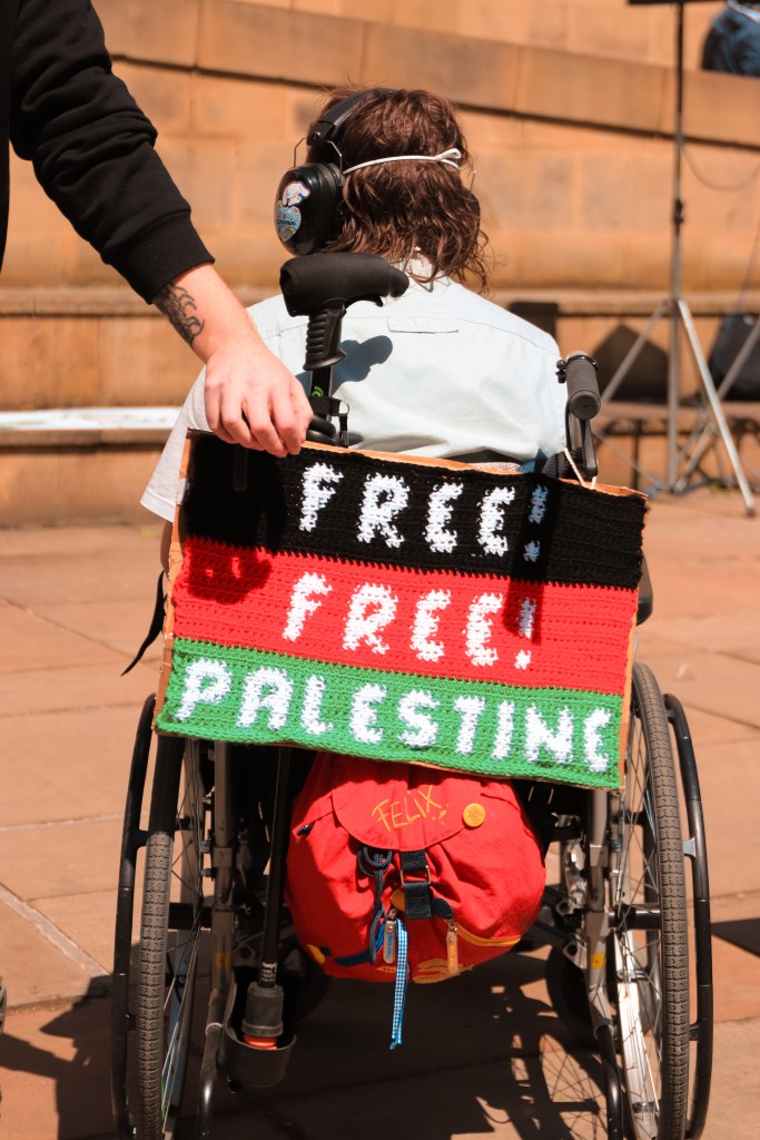 A crochet banner in black, red and green stripes, with 'Free! Free! Palestine' in white writing. The banner is hung on the back of a person's wheelchair, over their bag. The person in the wheelchair is facing away from the camera. They have brown wavy hair and are wearing ear defenders.
