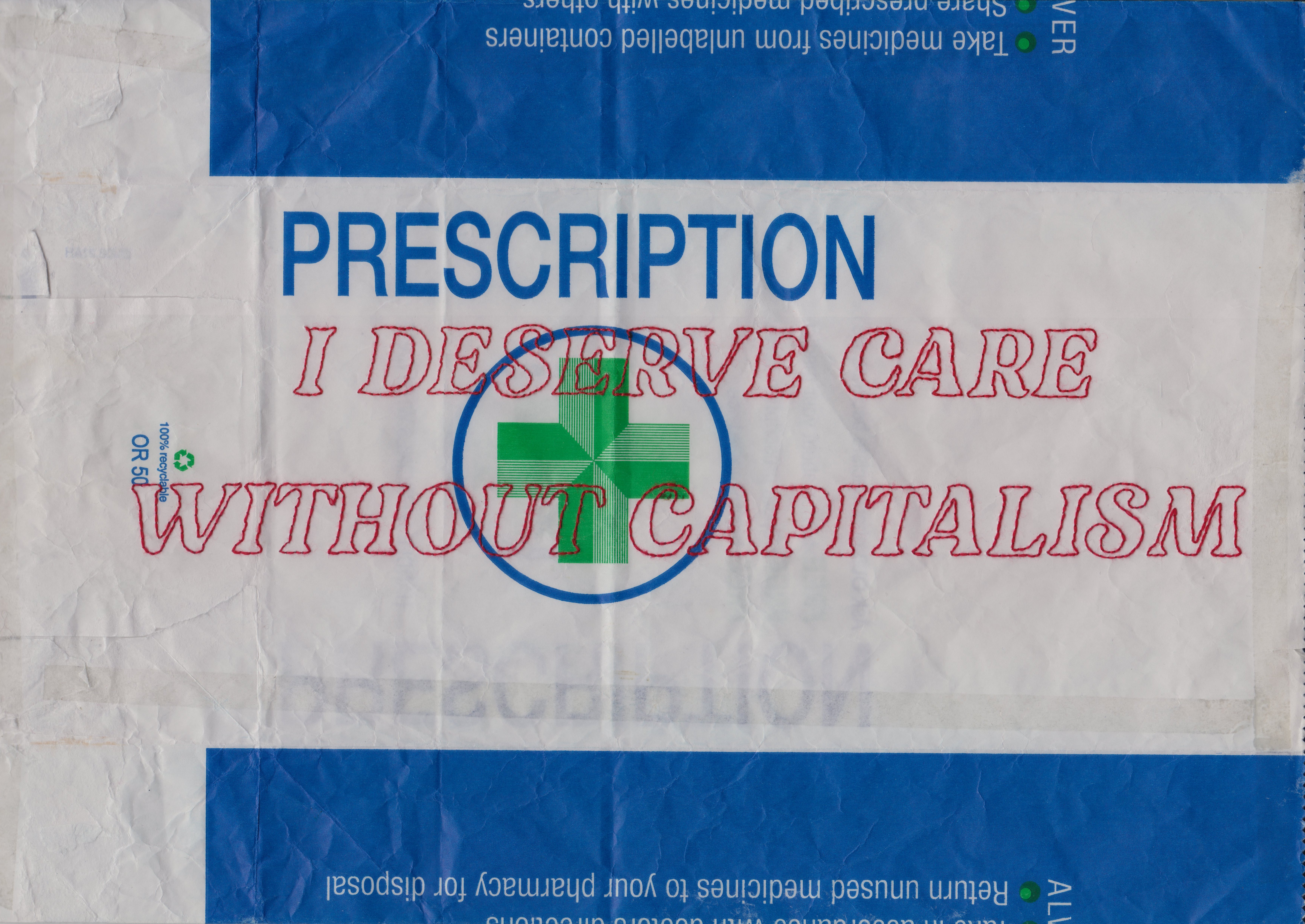 The phrase "I DESERVE CARE WITHOUT CAPITALISM" has been hand embroidered onto a paper pharmacy bag in bold capital letters using red thread and backstitch. It has been photographed against the backdrop of a mossy rock.