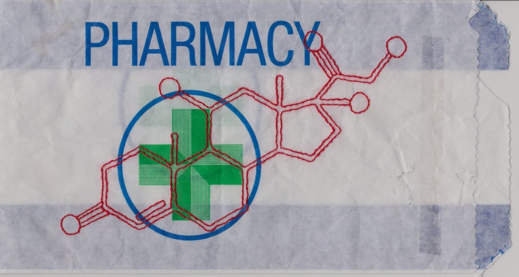 On a small paper pharmacy bag, the chemical symbol for the medication 'quetiapine' (made up of interconnecting hexagons, rectangles and circles), has been stitched in back stitch using red thread.