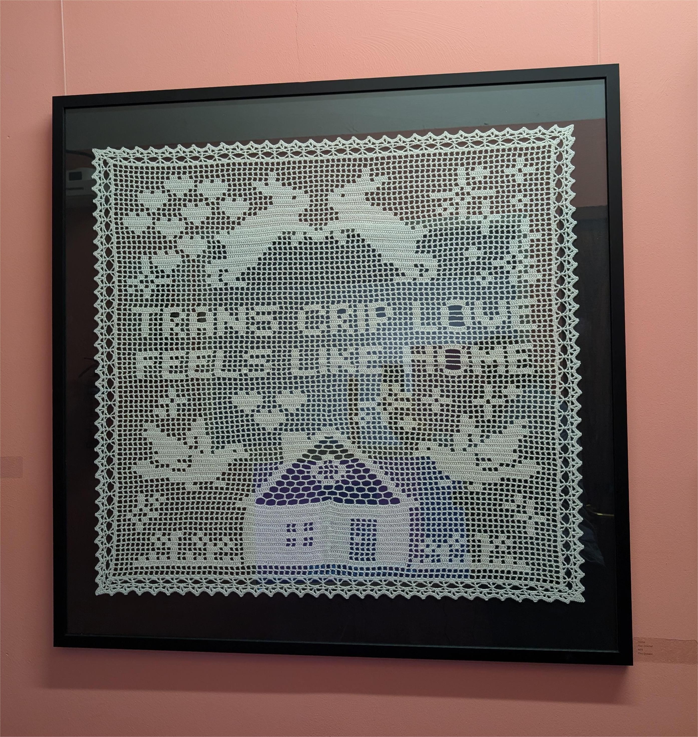 An image of the crochet piece 'Home' against a salmon pink wall, hung at exhibition