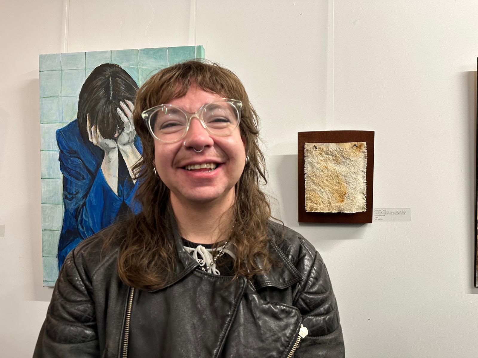 Finn (a white androgynous person with long, wavy, dark brown hair, large circular clear glasses, and nose piercings) stands in front of two pieces of art on a white gallery wall. They are wearing a black leather jacket and have a large grin on their face that scrunches up their eyes. To their left, slightly obscured, is a painting of a woman with her head in her hands by Jenny Meehan. To Finn's right is one of their own artworks: an orange textile piece nailed to a block of wood.