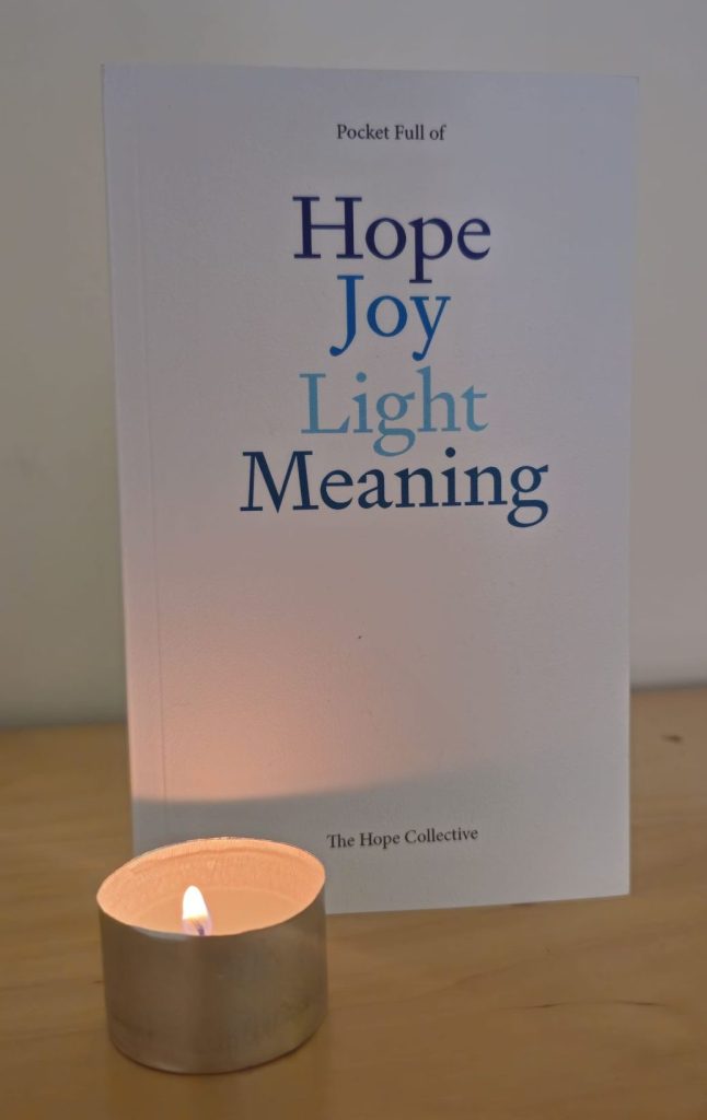 A book is stood upright on a shelf, lit by a tealight in the bottom left corner of the photo. The book cover is white, with the title: "Hope, Joy, Light, Meaning" laid out centrally in various shades of blue. In small text at the bottom are the collected authors: The Hope Collective.