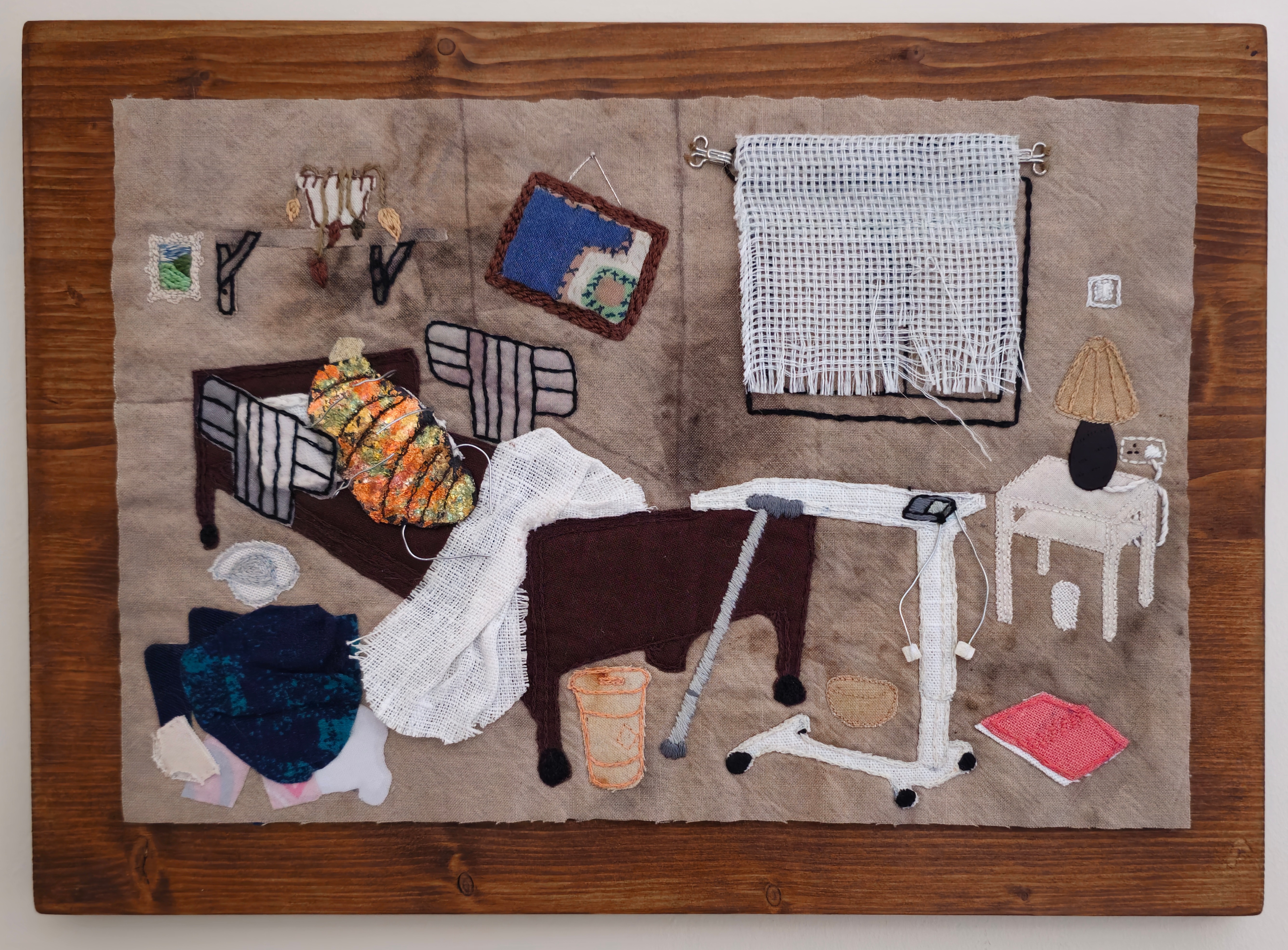 set against an oak-stained wood panel is a piece of textile art depicting a bedroom. It is in a state of dilapidation, with frayed curtains on the window, a framed picture hung at an angle, a dying plant on the shelf and clothes strewn on the floor. Central to the bedroom is a hospital bed, with barriers up at the sides, and laid in this is a bug, with iridescent gilding in oranges and greens, it's legs protruding out from the frame with the use of articulated wires. Various medical tools, such as a sharps bin, a tiny TENS machine, and a walking stick, are propped up around the bed. The scene feels vulnerable, as if peeking into someone's private domain. The use of the bug in the middle makes the viewer pause, and reconsider both the title and meaning behind the piece.
