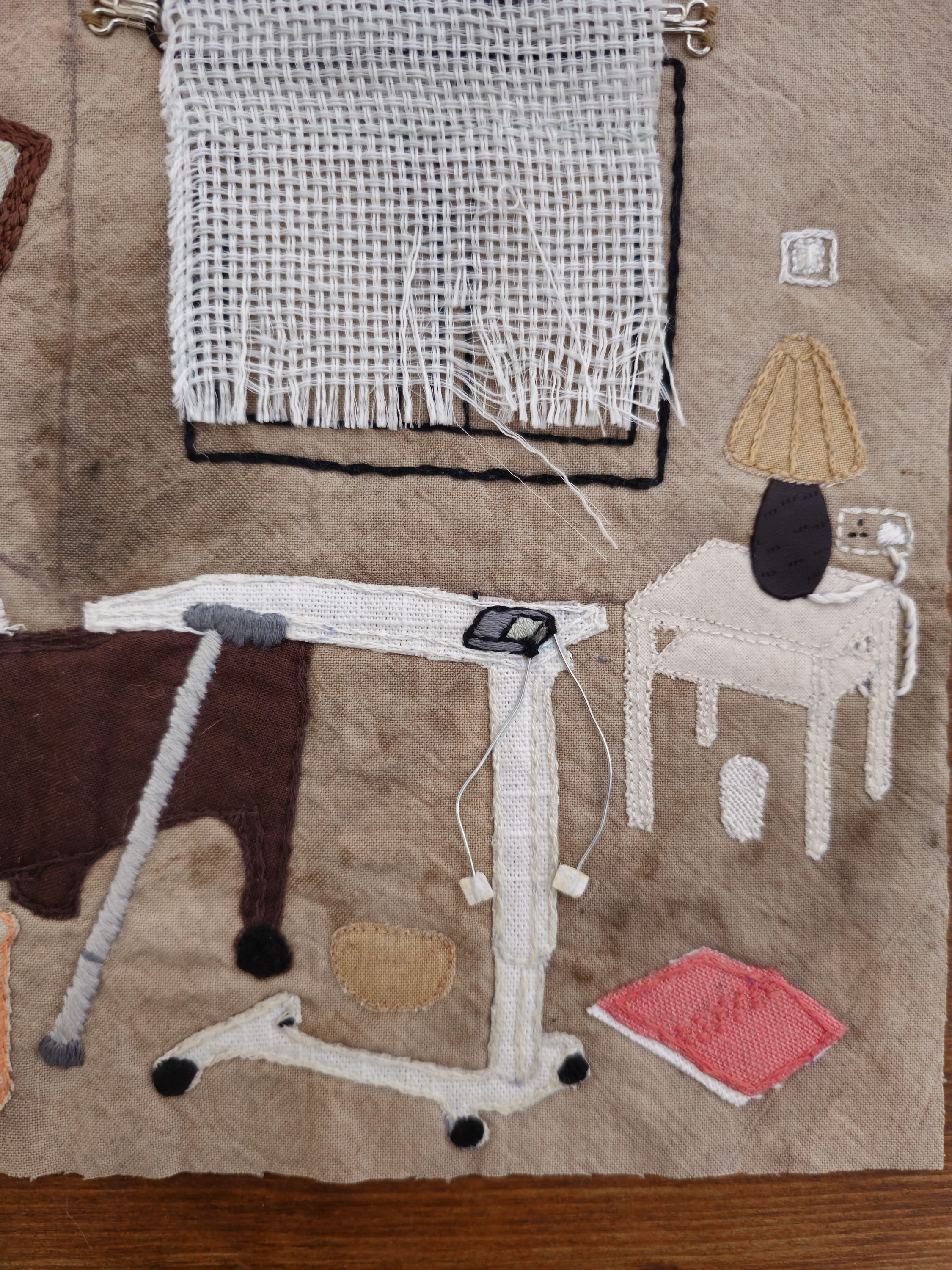 A close up of a piece of textile art, depicting a window covered in fraying curtains, a lamp on a side table, a hospital bed table, a TENS machine, a book lying open on the floor, and a walking stick propped against the table. The images have been created using fabric scraps, embroidery, wire and tiny scraps of paper.