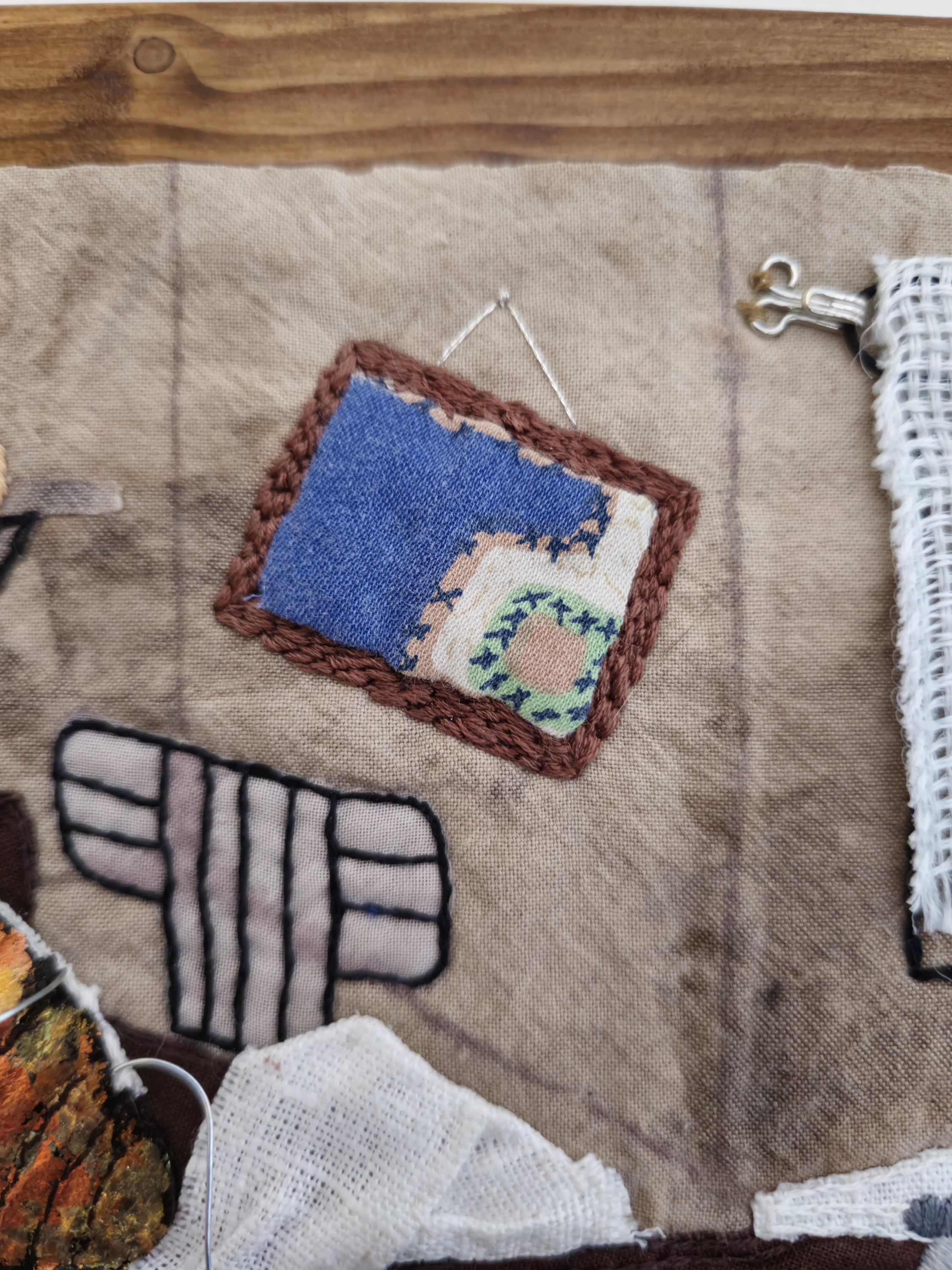 A close up of a piece of textile art. Depicted is a framed picture hung askew on a wall. The image is made up of a scrap of fabric, that has been carefully stitched around to create the frame.