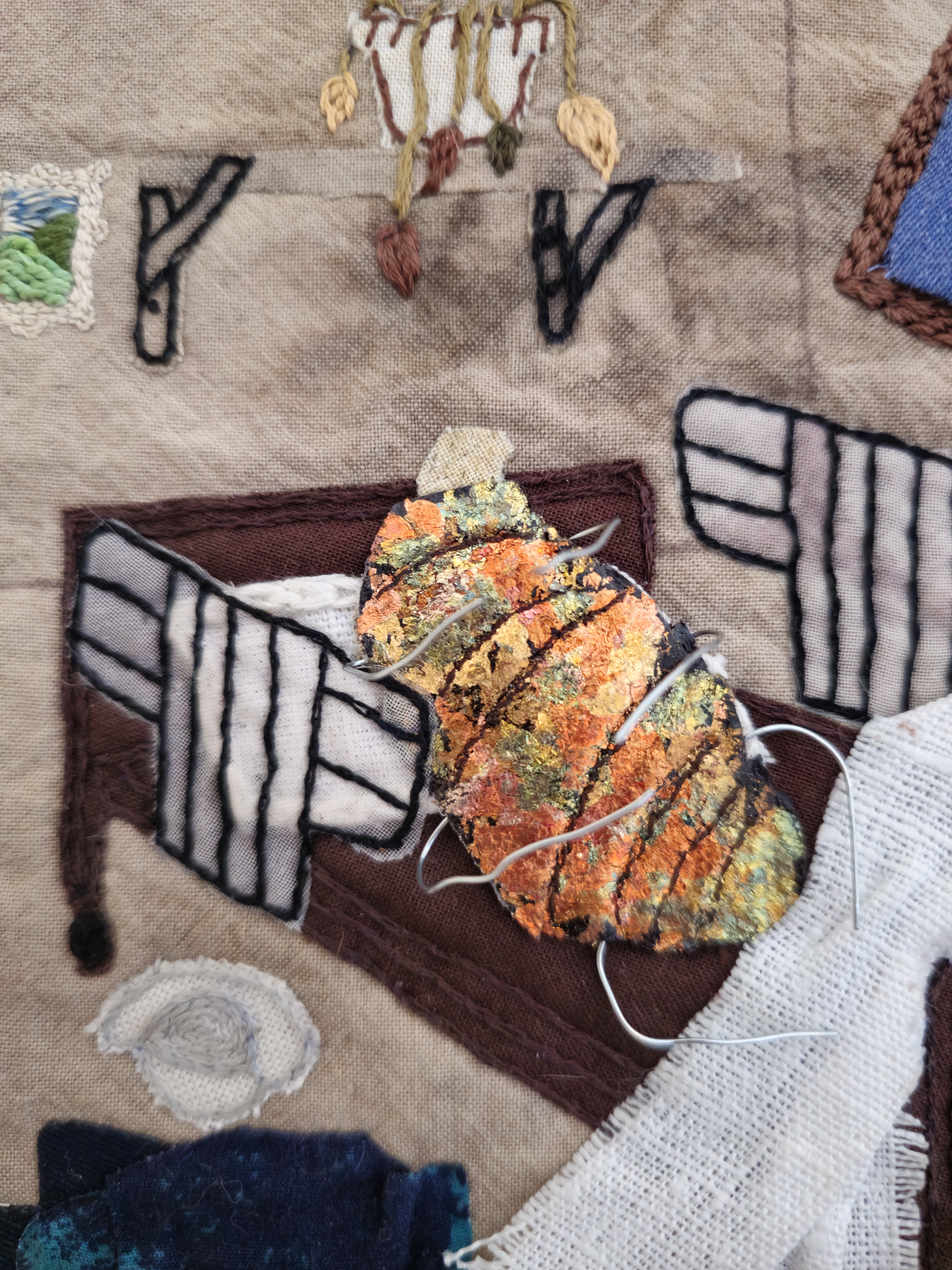 A close up of a piece of textile art that shows a bug in a hospital bed, surrounded by a sick bowl and dying plants. The bug's arms are made from wire, and have been articulated outwards to create a 3d effect. Its body is gilded with iridescent flakes in burnt orange, green and gold, reminding the viewer of a box of jewellery or chocolate wrappers. Embroidery stitches and details of fabric scraps are delicately used to make up the imagery