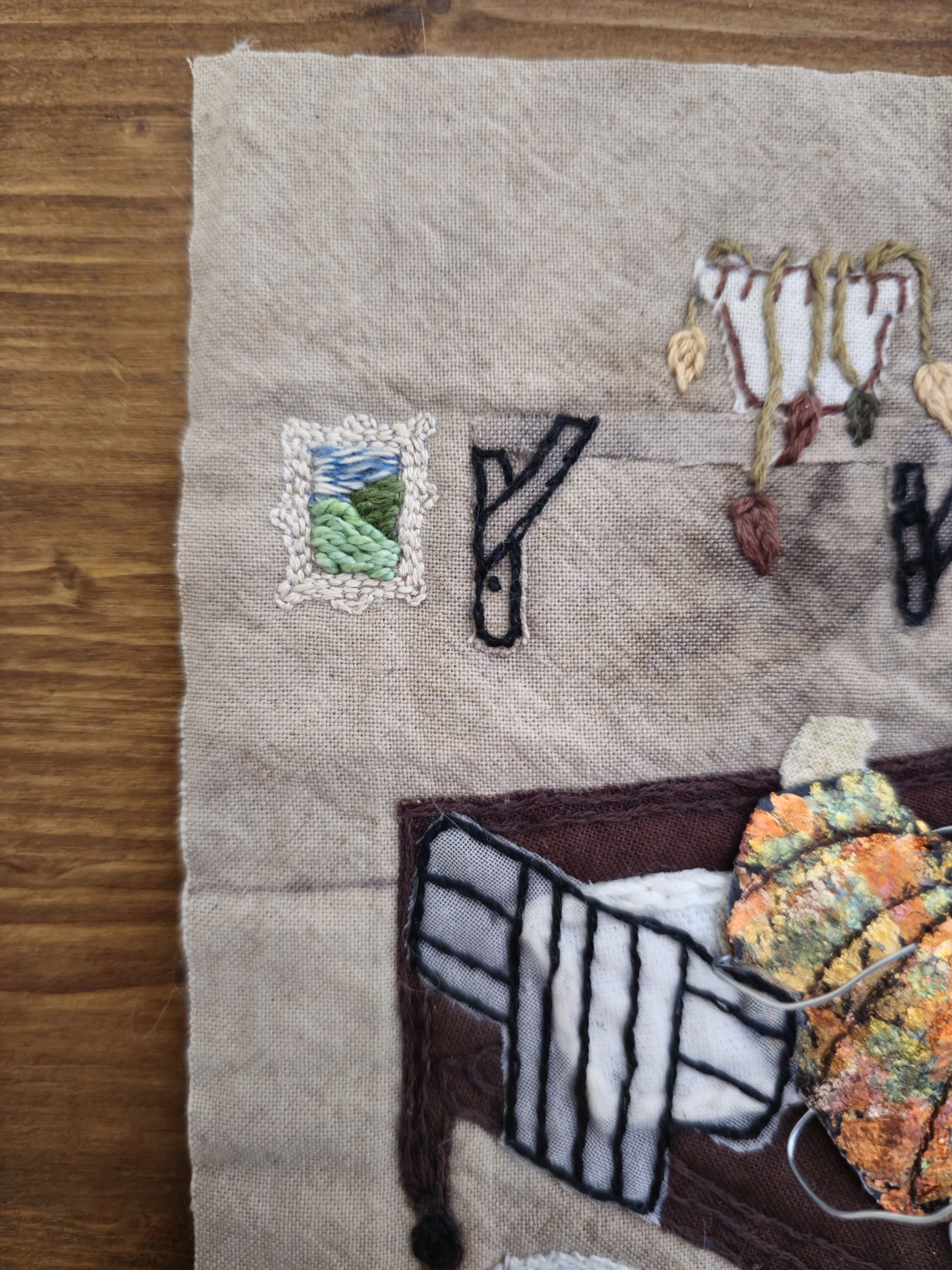 A close up of a piece of art that shows a framed images of hills that has been created using tiny stitches in cream, blue, white and shades of green. To the edge of the frame is a dying plant on a shelf, and a bug in a bed, created using fabric scraps and embroidery