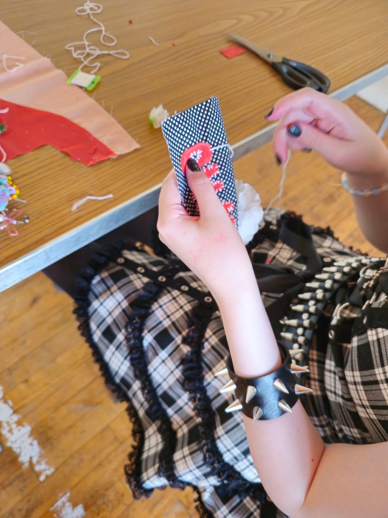 The camera  looks over the shoulder of a workshop participant, who has pale skin and is wearing a black and white plaid dress and gothic accessories. The picture captures their hands in motion as they stitch red cut out letters onto a black and white polka dot patch. In the background is a plastic faux-wood table with scissors, threads and fabric.