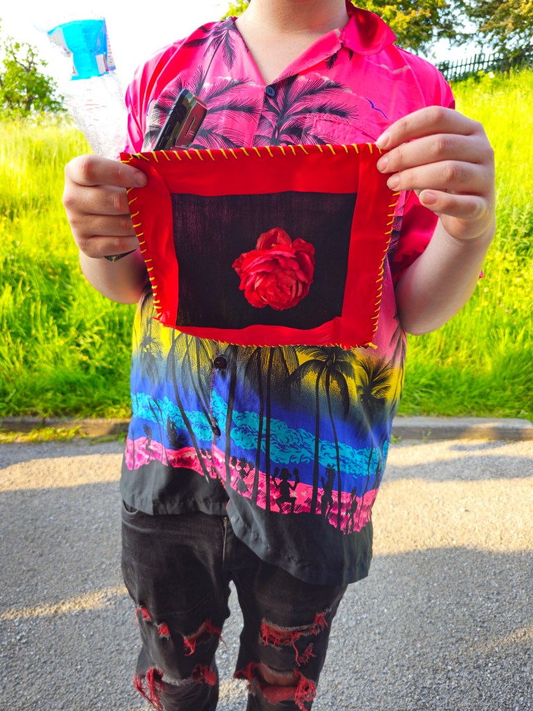 A workshop participant, their face out of frame, holds up their stitched patch to the camera. They are wearing a pink, yellow and blue Hawaiian shift and black trousers, and standing in a car park next to a field. The patch they are holding up is made of black fabric, with red ribbon stitched around all four sides, and a large red rose in the middle. 