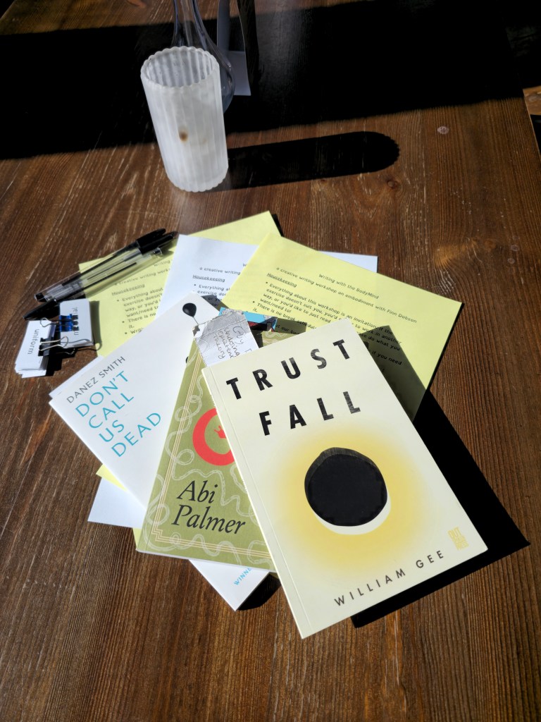 On a mahogany coloured table sit some workshop materials and glasses of water. The materials include workshop sheets on white and yellow paper, pens, and 3 books. These are, Trust Fall by Willam Gee, Slugs by Abi Palmer, and Don't Call Us Dead by Danez Smith
