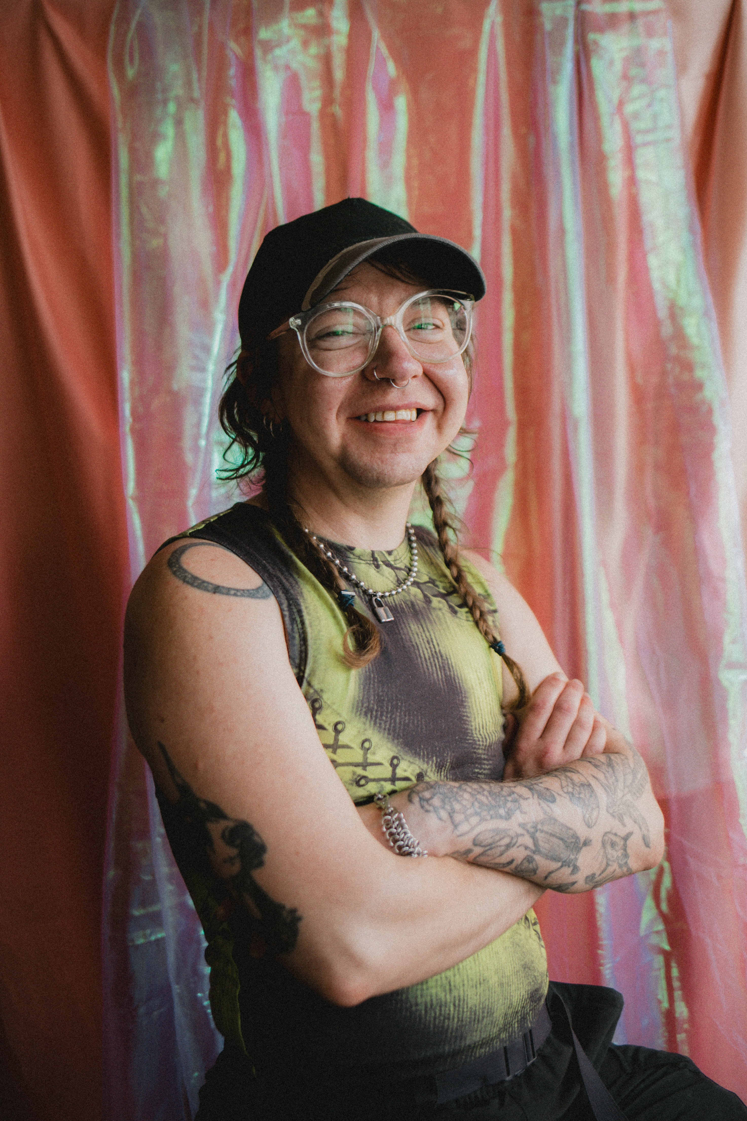 Finn, a white non-binary person with large clear glasses, nose piercings, and tattoos, is sat against a pink iridescent fabric background. They have their brown hair in long straggly plaits and are wearing a black cap and green and black tank top with stitching detailing. They're looking into the camera and smiling, with their arms crossed in front of them, in what appears to be a confident and happy demeanour.